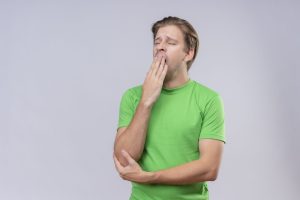 young handsome man wearing green t-shirt looking tired yawning standing over white background