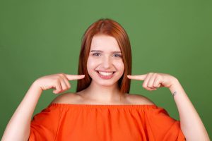 Young red hair woman in casual orange blouse on green background point fingers on white teeth perfect smile